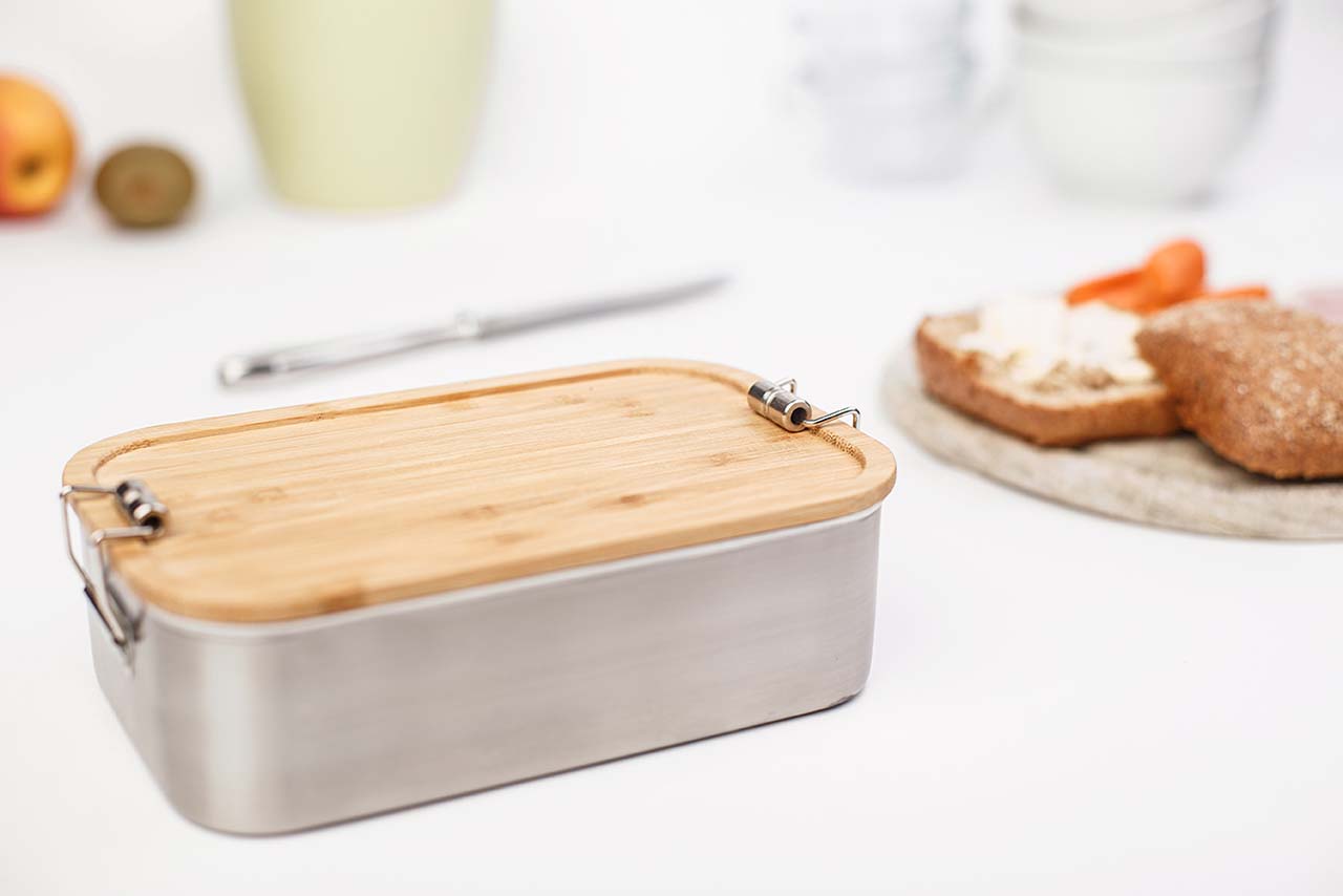 Rechteckige silberfarbene Metallbox mit abgerundeten Ecken und natürlichem Bambusdeckel, mit zwei seitlichen Verschlussklammern, auf einer weißen Küchenfläche