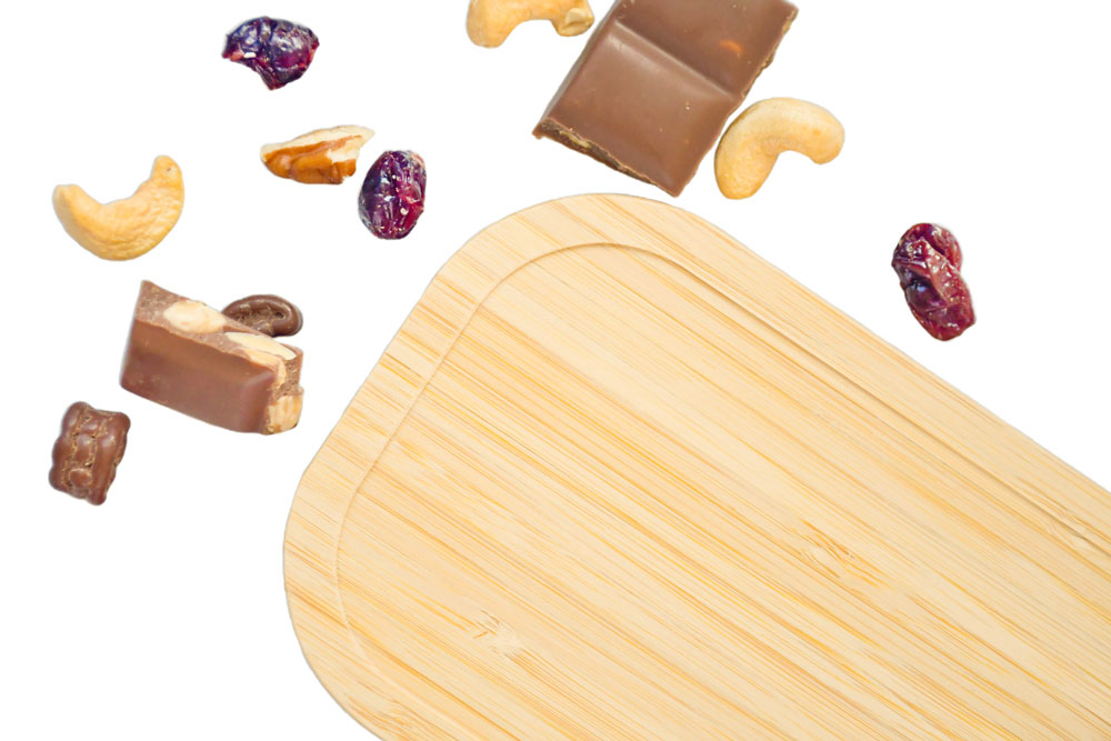 Bambusdeckel als Ersatzteil für Weißblech-Lunchbox, umgeben von Studentenfutter und Schokolade.